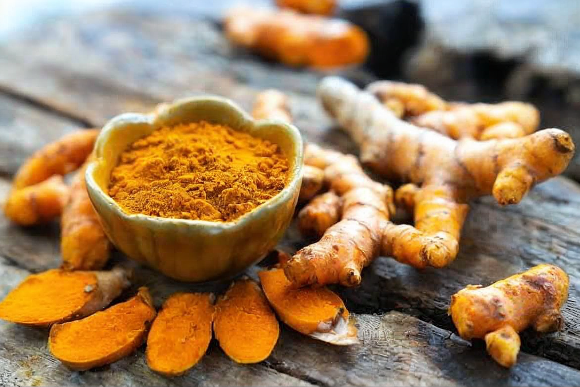 An earthy arrangement of turmeric (Curcuma longa) in its forms: a small bowl of golden turmeric powder surrounded by fresh, knobby turmeric root and bright orange slices, showcasing the natural spice as a holistic health and wellness ingredient.